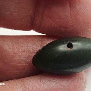 May include: A dark green, oval-shaped bead with a small hole. The bead is smooth and appears to be made of stone or a similar material. The object is held between fingers, suggesting it is a small, handheld item, possibly for jewelry or crafts.