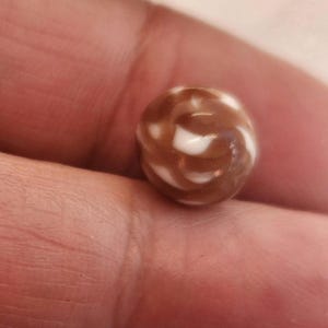 Puede incluir: Una canica marrón y blanca con remolinos. La canica tiene un acabado brillante y un patrón de remolinos marrones y blancos. Un juguete clásico, a menudo utilizado en juegos o como objeto de colección.