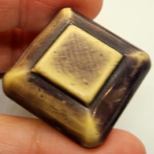 May include: A diamond-shaped button with a raised, square center. The button is made of a light brown material with a dark brown border. The center is a lighter shade of brown.
