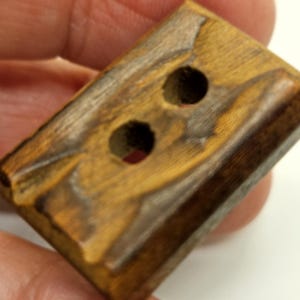 May include: A rectangular wooden button with two holes. The button has a natural wood grain pattern with shades of brown and tan. The edges are slightly beveled.