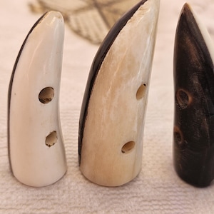 Puede incluir: Tres botones curvos en forma de cuerno, cada uno con dos agujeros. Los botones tienen una base de color crema con detalles en marrón oscuro o negro en los bordes. Los botones son probablemente para ropa o proyectos de artesanía.