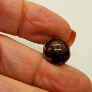 May include: A dark brown, round bead with a metallic gold edge. The bead is held between two fingers, showcasing its glossy surface and unique color variations. This bead could be used for jewelry making.