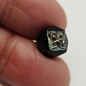 May include: A faceted, black button with a floral design. The button has a square shape with beveled edges. The floral design is iridescent with hints of green, gold, and purple. The button is held between two fingers.