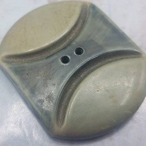 May include: A vintage, square button with a unique design. The button features a light gray, rounded top and a darker gray, diamond-shaped center. Two small holes are visible in the center for sewing. The button is made of a durable material.
