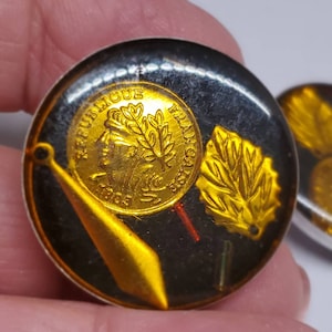 May include: Close-up of a pair of round, vintage earrings. Each earring features a gold-colored coin with the text "REPUBLIQUE FRANCAISE" and a gold leaf design set against a black background. The earrings have a metallic gold accent.