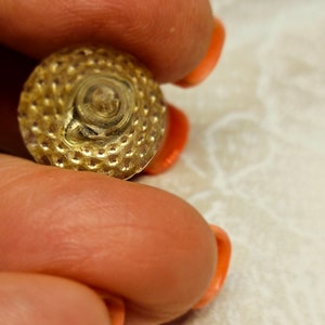 Puede incluir: Un botón de color dorado con un diseño circular texturizado. El botón tiene un patrón en espiral elevado en el centro.