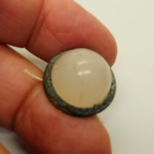 May include: A round, light pink button with a metallic, aged-looking border. A thin, off-white thread extends from the side. The button is held in a person's hand.