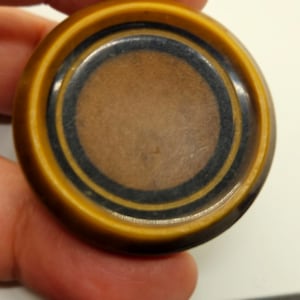 May include: A round, brown button with a black and gold concentric circle design.