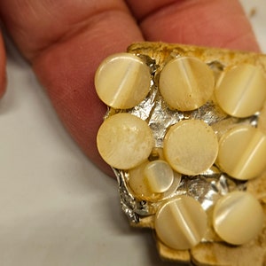 May include: Close-up of a collection of small, round, cream-colored buttons with a diagonal line across each. The buttons are attached to a piece of silver foil and a piece of cardboard.