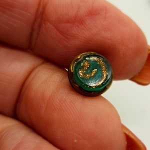 May include: A small, round earring with a dark green and gold design. The earring has a gold glitter pattern in the center and a dark brown border. The earring is held between fingers.