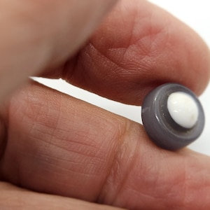 Puede incluir: Un pequeño botón redondo con un anillo exterior gris y un centro blanco. El botón se sostiene entre los dedos, mostrando su tamaño y diseño. Ideal para costura o proyectos de manualidades.