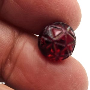 Puede incluir: Una cuenta facetada, redonda, de color rojo oscuro con un patrón geométrico. La cuenta se sostiene entre los dedos, mostrando su intrincado diseño y color. La superficie de la cuenta refleja la luz, resaltando sus facetas talladas.