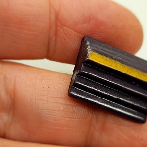 May include: A small, dark purple rectangular button with horizontal ridges and a yellow stripe. The button is held in a hand, showing its size and detail. The button's design is simple and geometric.