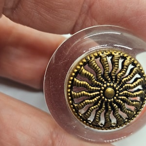 May include: A clear glass bottle stopper with a gold-toned metal design featuring a sunburst pattern. The design has a circular center with a smaller circle in the middle.