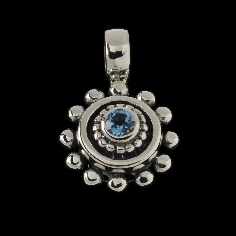 May include: A silver pendant with a blue gemstone in the center, surrounded by a circle of small beads. The pendant has a loop at the top for attaching a chain.