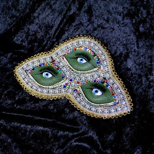 Puede incluir: Una máscara de carnaval con tres ojos verdes decorados con pedrería y cuentas de colores sobre un fondo de terciopelo negro.