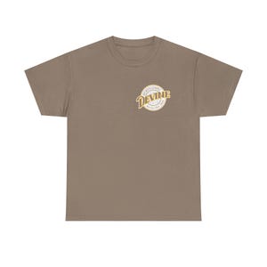 May include: A taupe-colored t-shirt with a crew neck. The shirt features a circular logo on the upper left chest with the words "Sports Gospel Devine" in a vintage style font. The logo is in shades of cream and gold.
