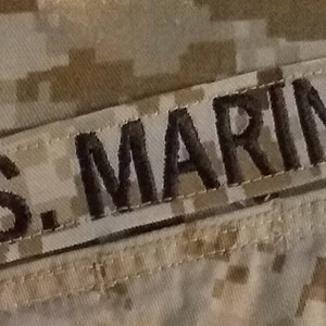 May include: A close-up of a military uniform patch with the text "U.S. MARINES" in black embroidery on a tan fabric background.