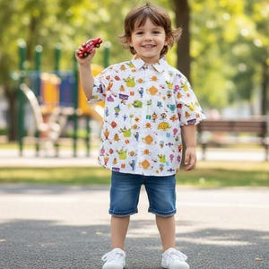 May include: A young child wearing a white button-down shirt covered in colorful cartoon monster illustrations, paired with denim shorts and white sneakers. The child is holding a small red toy car.