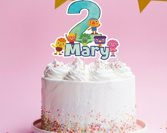 May include: A white cake with colorful sprinkles around the bottom. A cake topper with a blue number 2 and colorful cartoon characters says "Mary".