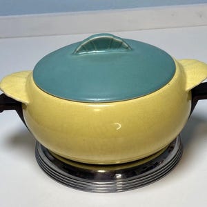 May include: A vintage yellow and teal ceramic casserole dish with a lid and handles. The dish sits on a chrome-coloured metal stand. The lid has a teal handle. The dish has a crackled glaze.