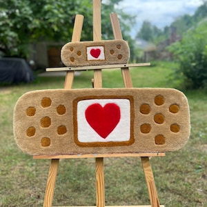 Może przedstawiać: Dwa dywany w kształcie plastra wystawione na drewnianym sztaludze. Większy dywan ma czerwone serce na białym kwadracie, otoczone brązową obwódką i brązowymi kropkami. Mniejszy dywan ma podobny wzór.