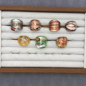 Puede incluir: Siete anillos de vidrio exhibidos en una bandeja de madera para joyas con ranuras de terciopelo blanco. Los anillos presentan varios patrones, incluyendo remolinos, rayas y diseños moteados en colores como plata, cobre, naranja, amarillo y verde.