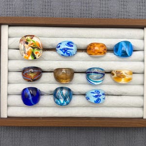 Könnte beinhalten: Eine Sammlung von elf Glasringen, die in einem Holztablett mit weißen Samtschlitzen präsentiert werden. Die Ringe weisen verschiedene Muster und Farben auf, darunter marmorierte blaue, orangefarbene, braune und mehrfarbige Wirbel.