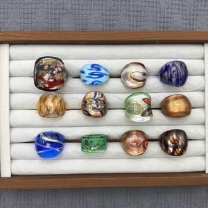 Op de afbeelding: Een collectie van twaalf glazen ringen gerangschikt in een houten presentatieblad met witte fluwelen sleuven. De ringen hebben verschillende patronen en kleuren, waaronder blauw, groen, goud, bruin en zwart, met wervelende, metallic en gespikkelde ontwerpen.