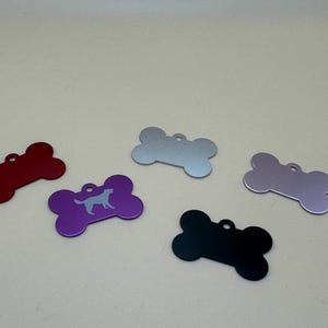 Peut inclure: Cinq médailles d'identification pour animaux en forme d'os de différentes couleurs : rouge, violet, argenté, violet clair et noir. La médaille violette présente une silhouette de chien claire. Chaque médaille a un petit trou circulaire.