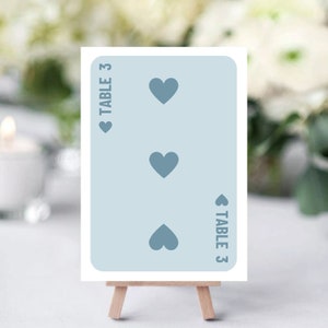 May include: A light blue card with three heart symbols and the text "TABLE 3" on a small wooden easel. The card is a table number for an event.