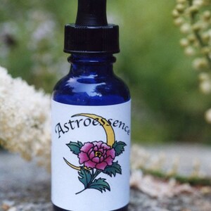 Puede incluir: Una botella de vidrio azul con un gotero y una etiqueta blanca. La etiqueta tiene una luna creciente amarilla y una flor rosa con hojas verdes. El texto de la etiqueta dice "Astroessence".