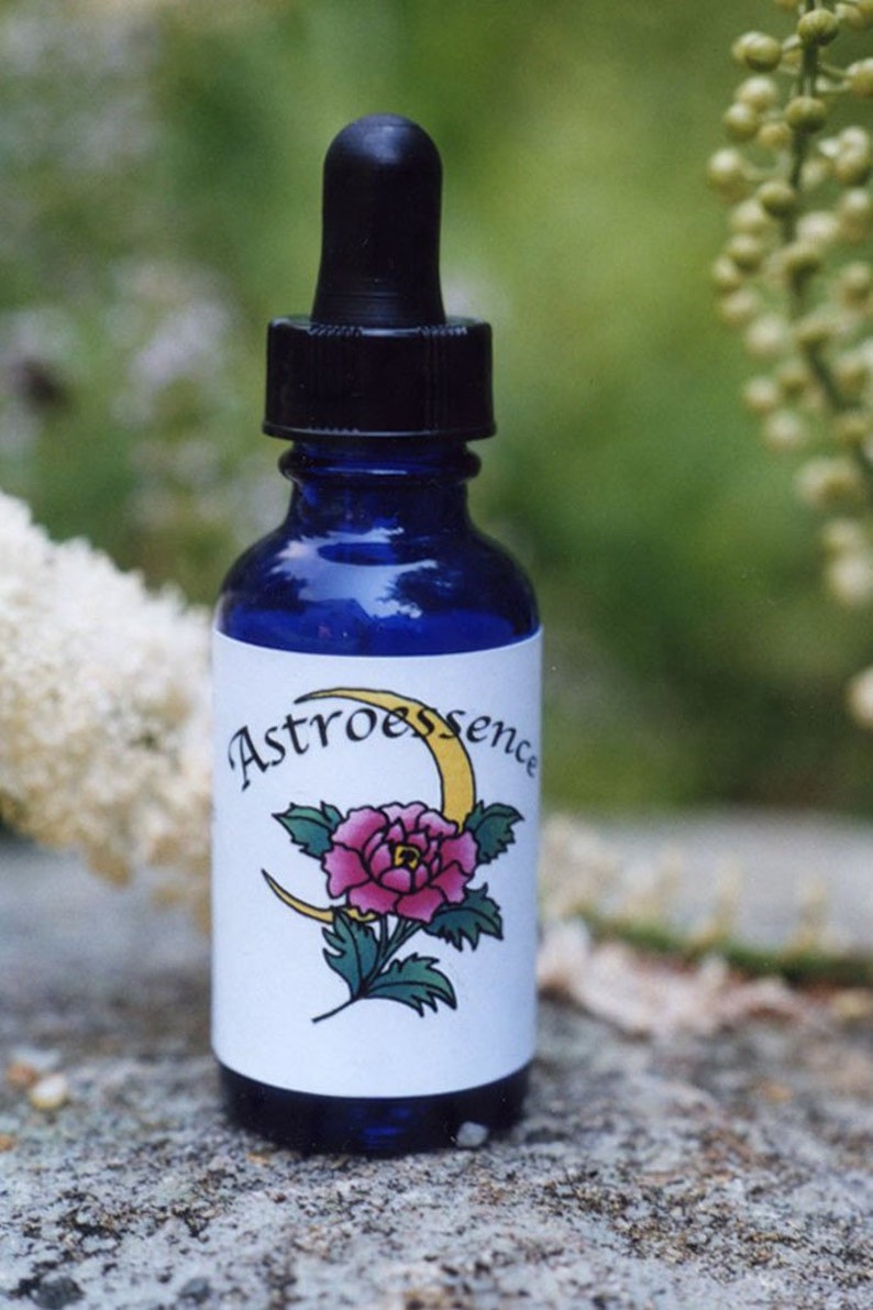 Puede incluir: Una botella de vidrio azul con un gotero y una etiqueta blanca. La etiqueta tiene una luna creciente amarilla y una flor rosa con hojas verdes. El texto de la etiqueta dice "Astroessence".