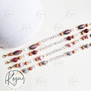 May include: Four sports-themed beaded lanyards with gold clasps. The lanyards feature football, basketball, baseball, and soccer beads, along with wooden and white beads. The words "FOOTBALL", "BASKETBALL", "BASEBALL", and "SOCCER" are spelled out in white letter beads.