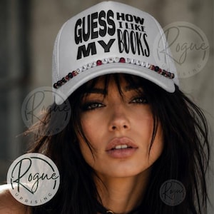 Może przedstawiać: Biała czapka typu trucker z czarnym napisem "GUESS MY BOOKS HOW I LIKE". Rondo zdobią koraliki z napisem "DARK AND DIRTY". Czapka ma siatkową tylną część.