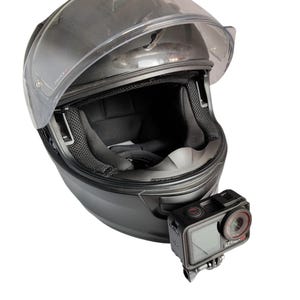 Peut inclure: Casque de moto noir avec une visière transparente et une caméra d'action noire fixée. Le casque a un intérieur rembourré et un design épuré. La caméra d'action est noire avec un objectif rouge.
