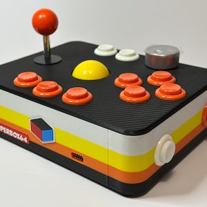 Może przedstawiać: Kontroler do gier Superbox64 w stylu retro z czarnym blatem i kolorowymi paskami bocznymi. Posiada pomarańczowy joystick, pomarańczowe i białe przyciski oraz żółty środkowy przycisk. Widoczny jest tekst "SUPERBOX64".
