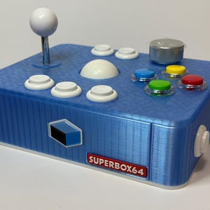 Puede incluir: Un mando de juegos Superbox64 azul con un joystick y botones blancos. El mando tiene una variedad de botones de colores, incluyendo azul, amarillo, verde y rojo. El diseño incluye un joystick blanco en forma de bola y una perilla plateada.