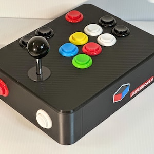 Op de afbeelding: Zwarte arcadecontroller met een koolstofvezeltextuur, met een joystick en kleurrijke knoppen. De controller heeft rode, witte, gele, blauwe en groene knoppen. De tekst "SUPERBOX64" is zichtbaar aan de zijkant.