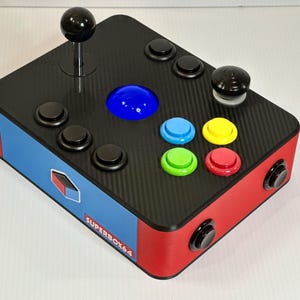 以下が含まれることがあります： 黒と赤のアーケードゲームコントローラー。カーボンファイバーの質感で、ジョイスティック、いくつかの黒いボタン、青、緑、黄色、赤のカラーボタンを備えています。コントローラーの側面には「SUPERBOX64」の文字があります。