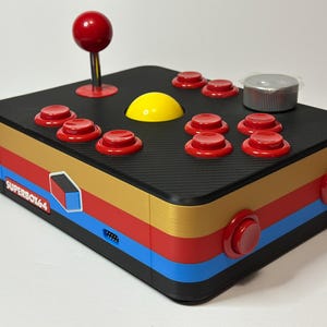Puede incluir: Un mando de juego retro negro con botones rojos y un joystick. El mando tiene un botón amarillo y un dial plateado. El lateral del mando tiene un diseño de rayas azules, rojas y doradas. El texto "SUPERBOX64" es visible.