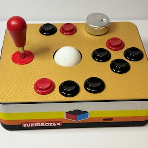 Op de afbeelding: Een retro Superbox64 gamecontroller met een gele, getextureerde bovenkant, een rode joystick en diverse zwarte en rode knoppen. Het heeft een zilveren knop en een witte bal. De basis heeft een gestreept ontwerp met de tekst "SUPERBOX64".
