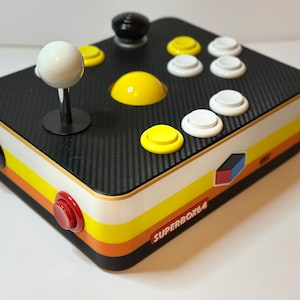Könnte beinhalten: Ein Arcade-Gamecontroller im Retro-Stil mit einer schwarzen, mit Kohlefaser strukturierten Oberseite. Er verfügt über einen weißen Joystick, gelbe und weiße Knöpfe sowie einen roten Knopf. Die Seiten haben gelbe, orange und weiße Streifen. Der Text "SUPERBOX64" ist sichtbar.