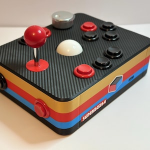 Może przedstawiać: Kontroler do gier Superbox64 w stylu retro z czarnym wierzchem z włókna węglowego i kolorowymi paskami bocznymi. Posiada czerwony joystick, czerwone i czarne przyciski oraz srebrny przycisk. Widoczny jest tekst "SUPERBOX64".