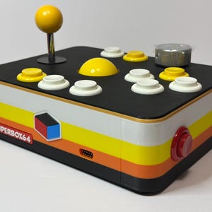 Może przedstawiać: Kontroler do gier zręcznościowych Superbox64 w stylu retro. Posiada czarny wierzch z żółtym joystickiem i przyciskami oraz białymi i żółtymi przyciskami. Boki są białe z żółtymi, pomarańczowymi i czarnymi paskami. Posiada czerwony i czarny przycisk.