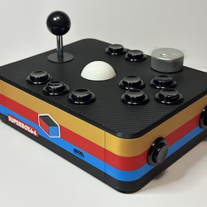 Puede incluir: Controlador de arcade Superbox64 negro y dorado con un joystick y botones negros. El controlador tiene una textura de fibra de carbono en la parte superior, una bola blanca y una perilla plateada. Los lados presentan rayas rojas, doradas y azules.