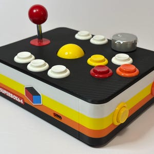 Può includere: Un controller arcade Superbox64 in stile retrò con una parte superiore nera e strisce laterali colorate. È dotato di un joystick rosso, vari pulsanti colorati e un quadrante argentato. Il design ricorda le console di gioco classiche.