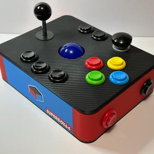 以下が含まれることがあります： 黒いカーボンファイバーテクスチャのトップ、赤と青の側面、黒いジョイスティックを備えたSuperbox64アーケードスタイルのコントローラー。黒、赤、黄、緑、青のボタンがあります。「SUPERBOX64」の文字が青い側面に見えます。