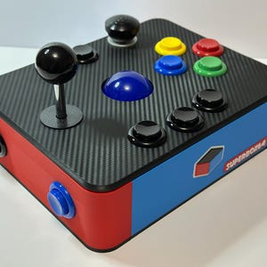 以下が含まれることがあります： 赤、青、黒のアーケードゲームコントローラー。カーボンファイバーのトップ、ジョイスティック、黒いボタン、カラフルなボタンが特徴です。「SUPERBOX64」の文字が側面に表示されています。