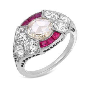 May include: A platinum ring featuring a central round rose-cut diamond surrounded by a cluster of smaller round diamonds and two curved rows of square-cut vibrant red rubies. The metal band displays intricate milgrain detailing along the edges.